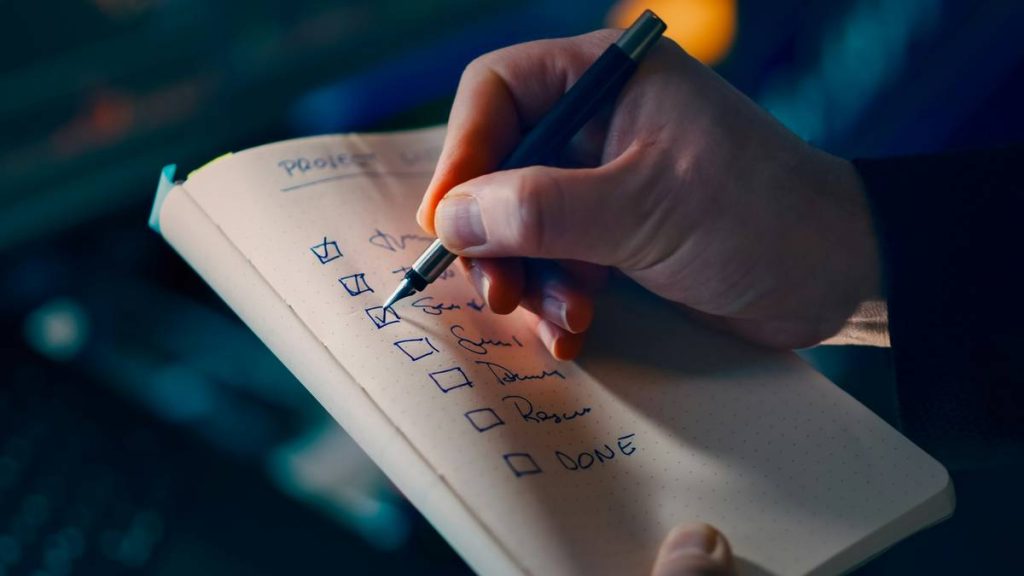 Project checklist planning productivity concept. Closeup hand writing tasks notebook pen focus workflow organization time management business efficiency workplace routine